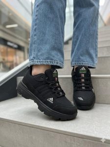 نمای نزدیک از کفش کتانی زنانه مشکی آدیداس (Adidas) پوشیده شده با شلوار جین روی پله‌ها.